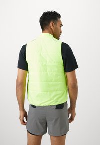 Gilet imbottito giallo brillante con design trapuntato, senza maniche e con dettagli riflettenti. Indossato sopra una maglietta nera e pantaloni corti grigi.