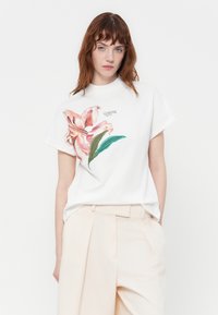 T-shirt bianco in cotone con colorata ricamo floreale sul lato sinistro, impreziosito da un sottile logo del marchio. Maniche corte e vestibilità rilassata.