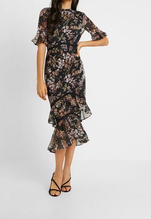 Femme portant une robe noire à motif floral avec un ourlet et des manches volantés, associée à des sandales noires à talons hauts à lanières, se tenant devant un fond uni.