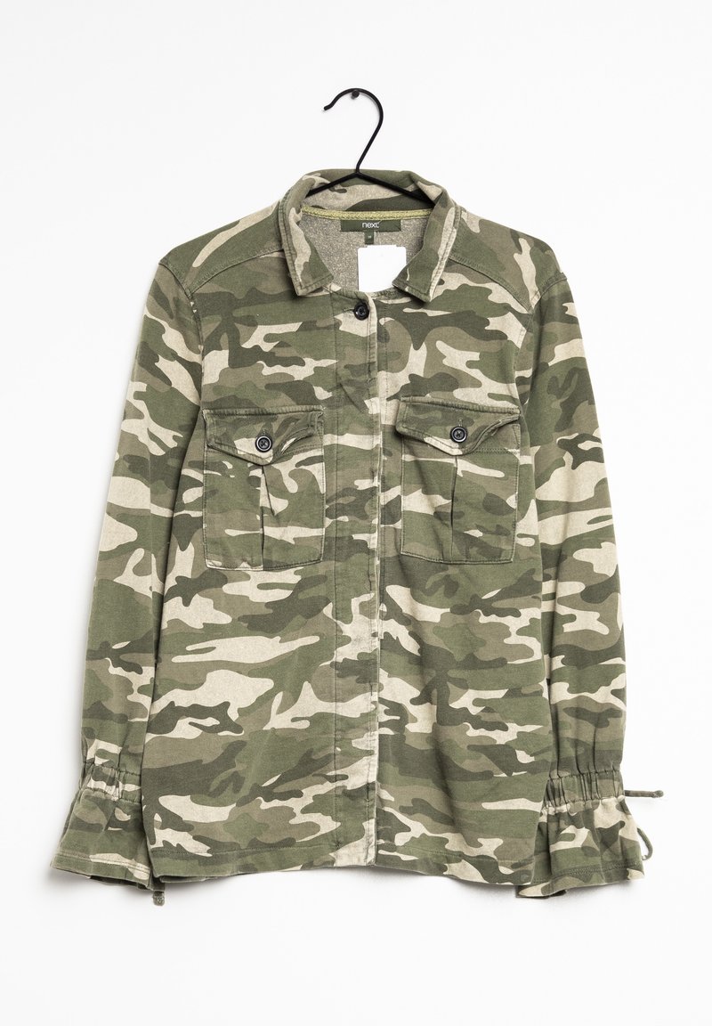 Chemise boutonnée camouflage verte et beige avec deux poches poitrine, manches longues avec poignets volantés ajustables, suspendue sur un cintre noir.