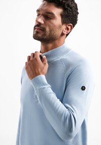 Lichtblauw poloshirt met lange mouwen, voorzien van een ritskraag en ribbels aan de mouwen. Heeft een zwart hexagonaal logo op de linkermouw.