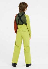 Maglia a maniche lunghe verde e arancione a strati abbinata a pantaloni da neve giallo vivace con bretelle regolabili e tasche laterali. Stivali neri visibili.