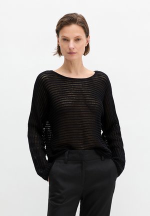 Frau mit kurzen braunen Haaren trägt einen schwarzen, offen gestrickten Pullover mit langen Ärmeln und schwarze, hoch taillierte Hosen, steht mit den Händen in den Taschen.