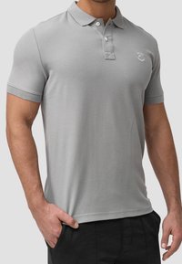 Polo shirt gris clair en tissu tricoté. Il présente un col, une patte de boutonnage à deux boutons et un petit logo sur la poitrine. Manches courtes.