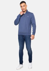Hombre con un suéter azul de media cremallera, pantalones ajustados oscuros azul marino y zapatillas blancas, de pie contra un fondo claro y liso.