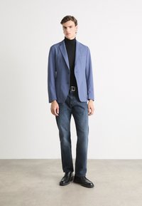 Niebieska marynarka na czarnej golfie, w połączeniu z ciemnymi dżinsami i czarnymi butami. Marynarka ma slim fit i klapy z wycięciem.