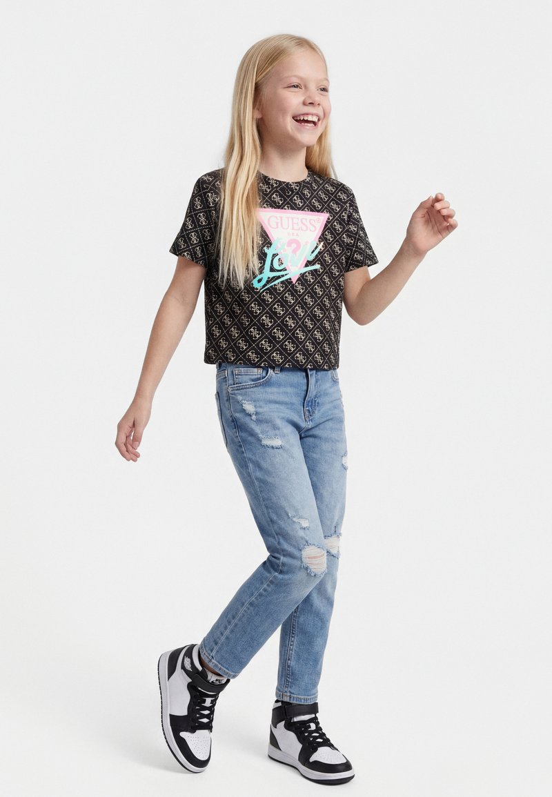 Fată tânără zâmbitoare, mergând, purtând un tricou negru cu model Guess Love, blugi albaștri rupți și adidași alb-negru pe fundal alb.