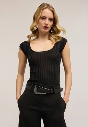 Blondynka w dopasowanym czarnym prążkowanym topie z krótkim rękawem, czarnych spodniach z szerokim czarnym paskiem, ręce w kieszeniach, neutralne tło.