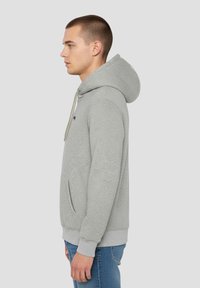 Sweat à capuche gris clair avec poche kangourou, capuche avec cordon de serrage et poignets côtelés. Tissu texturé légèrement brillant, porté avec un jean bleu.