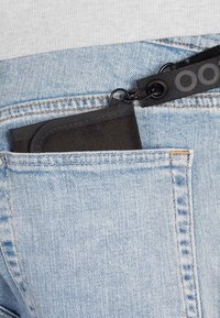 Black fabric cardholder partially visible in a light blue denim pocket, secured with a black strap and metal clip, featuring rounded edges.