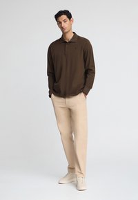 Brun långärmad pikétröja med krage och tre knappar, matchad med beiga byxor och ljusa sneakers. Enkelt, figursytt design.