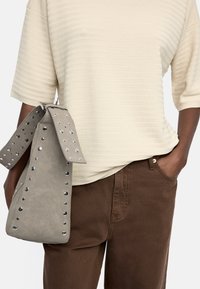 Szara zamszowa torba z ćwiekami i szerokim paskiem. Nosi się ją z beżową bluzką w paski i brązowymi spodniami, co podkreśla warstwowy strój.