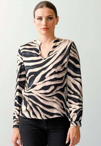 Frau trägt eine langärmlige Bluse im Zebra-Muster mit einem kleinen V-Ausschnitt und schwarze Hosen und steht vor einer schlichten hellen Wand.