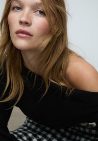 Crna pulover bluza bez ramena s mekim teksturom, u kombinaciji s crno-bijelim kariranim suknjom. Prirodna, neuređena boja kože.