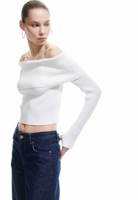 Valkoinen off-the-shoulder ribattu crop-top, jossa on pitkät hihat, yhdistettynä korkeavyötäröisiin tummansinisiin farkkuihin. Sujuva pinta ja tiukka muotoilu.