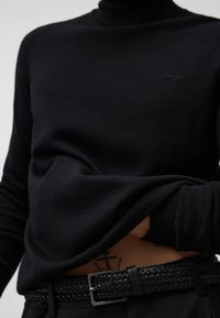 Czarny sweter z wysokim kołnierzem w gładkim materiale, zestawiony z plecionym czarnym paskiem z prostokątną metalową klamrą, widoczne detale tatuażu poniżej.