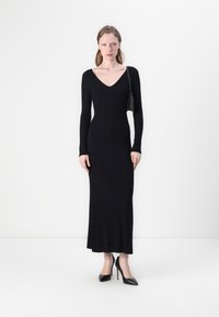 Black long-sleeve dress with a V-neck, ribbed texture, fitted silhouette, and floor-length design. Model accessorized with a small bag.