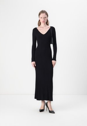 Black long-sleeve dress with a V-neck, ribbed texture, fitted silhouette, and floor-length design. Model accessorized with a small bag.