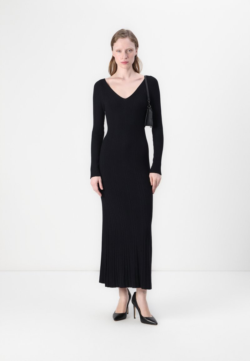 Black long-sleeve dress with a V-neck, ribbed texture, fitted silhouette, and floor-length design. Model accessorized with a small bag.