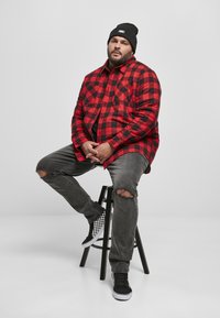 Rød og svart rutete flanellskjorte, grå slitte jeans, og svart og hvit rutete joggesko, båret med en svart lue, sittende på en krakk.
