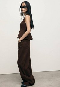 Brown peplum top paired with wide-leg brown trousers. Model wears dark sunglasses and black footwear. Clean, minimalist design.