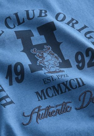 Zils kokvilnas T-krekls ar centrālo logo, kurā attēlots briedis, un tekstu "H Club Original 1992" un "Authentic Decor" kontrastējošās melnās un brūnās krāsās.