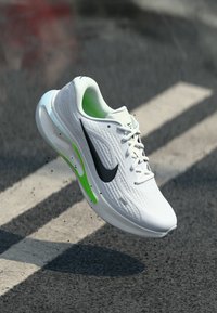 Άσπρο αθλητικό παπούτσι με υφή, μαύρο λογότυπο swoosh, πράσινες λεπτομέρειες και ελαφρύ σχεδιασμό. Αντικείμενο σε επίπεδη επιφάνεια.