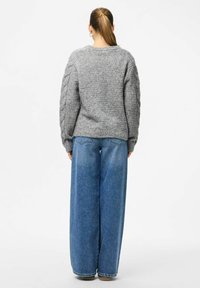 Femme avec une queue de cheval portant un pull en tricot gris et un pantalon en jean bleu à large jambe, se tenant de dos contre un fond blanc.