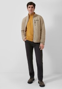 Mann trägt eine beige Fleecejacke mit Reißverschluss über einem senfgelben Pullover, schwarze Hose und braune Schnürschuhe, steht vor einem schlichten, hellen Hintergrund.