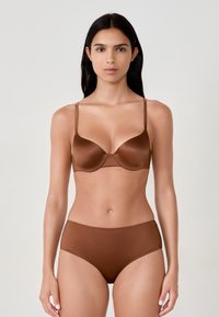 Donna con lunghi capelli scuri, che indossa un reggiseno di satin marrone e mutandine senza cuciture coordinate, in piedi contro uno sfondo chiaro e semplice.