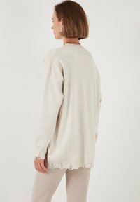 Maglione lavorato a maglia beige chiaro con vestibilità ampia, polsini a coste e orlo a scallop con dettagli sfrangiati. Texture morbida, maniche lunghe.