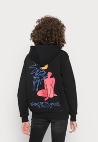 Hoodie preto com uma gráfica colorida nas costas: uma figura sentada em rosa, folhas de palma azuis e o texto "FÉMINISTE TOUJOURS." Textura suave.