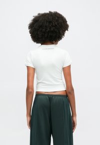 Balts, pieguļošs īsām piedurknēm t-krekls ar apaļu kakla izgriezumu un "NIKE SHOX" logo tuvu kaklam, apvienots ar tumši zaļām, brīvi pieguļošām biksēm.