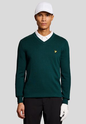 Man met witte pet, donkergroene V-hals trui met gele vogel logo, wit poloshirt, zwarte broek en witte golfhandschoen aan de rechterhand.