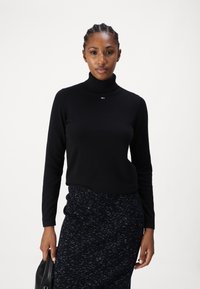 Melnas turtlenecka kreklas ar garām piedurknēm, izgatavots no mīksta materiāla. Uz krūšu daļas neliels logo akcents. Apvienots ar rakstainām tumšām svārkiem.