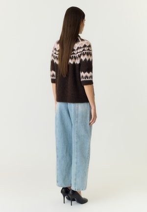 Femme aux longs cheveux bruns portant un pull à motifs marron foncé et rose, un jean ample bleu clair et des chaussures à talons noirs, debout, de dos.