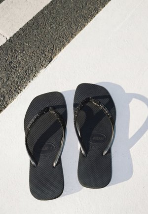 Paar schwarze Havaianas Flip-Flops mit dünnen Riemen, die auf einer weißen Oberfläche nahe dem strukturierten Asphaltrand liegen.
