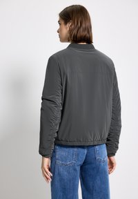 Giacca bomber grigia con colletto a coste e polsini elasticizzati, caratterizzata da maniche testurizzate e orlo aderente, abbinata a jeans in denim blu.