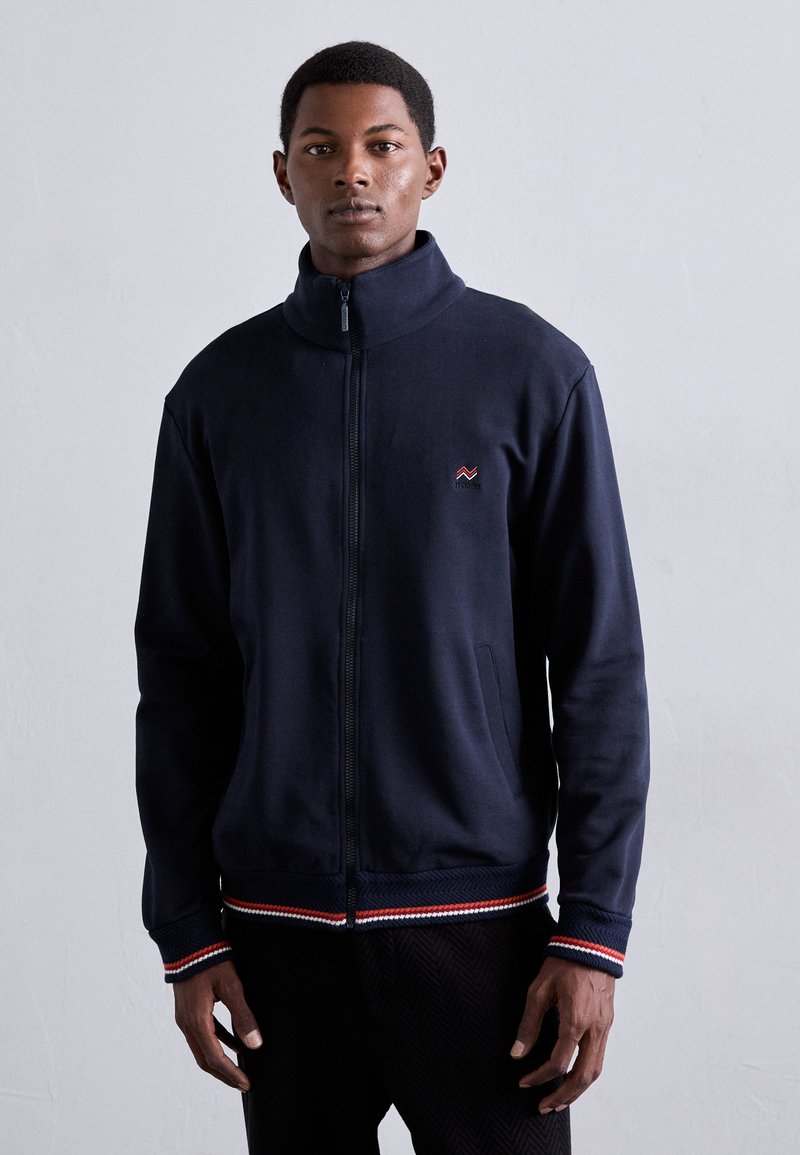 Marinefarbene Zip-Jacke aus weichem Material mit hohem Kragen, seitlichen Taschen und einem gestreiften Saum in Rot- und Weißakzenten.