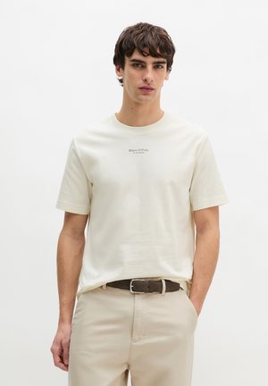 Jonge man draagt een off-white Marc O'Polo T-shirt, ingestopt in beige broek met een bruine riem, staande tegen een effen witte achtergrond.