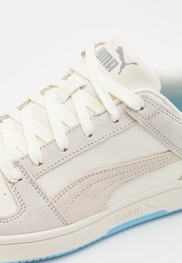 Sneakers Puma z kremową skórzaną cholewką i jasnoszarymi zamszowymi akcentami. Posiadają białe sznurówki, teksturowaną podeszwę środkową i niebieską podeszwę zewnętrzną.