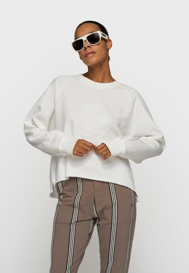 Mujer con sudadera blanca de gran tamaño con letras en relieve, pantalones marrones a rayas y grandes gafas de sol blancas, posando frente a un fondo liso.