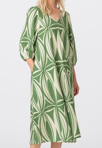 Green and cream patterned dress with a V-neck, puffed sleeves, and side pockets; features a floral motif and relaxed silhouette.