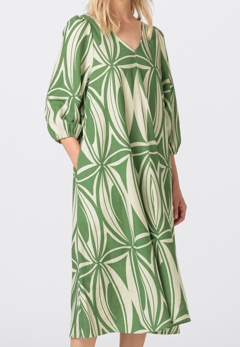 Green and cream patterned dress with a V-neck, puffed sleeves, and side pockets; features a floral motif and relaxed silhouette.