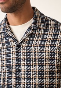 Chemise à manches courtes présentant un motif tissé bleu marine, blanc et marron, avec un col en encoche et des boutons noirs sur un tissu texturé.