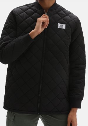 Person trägt eine schwarze gesteppte Vans-Jacke mit Frontreißverschluss und hält den Reißverschlussgriff nahe am Kragen.