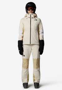 Abbigliamento da sci con una palette di colori crema e beige, giacca impermeabile con tasche con zip e pantaloni isolati con ginocchia rinforzate.