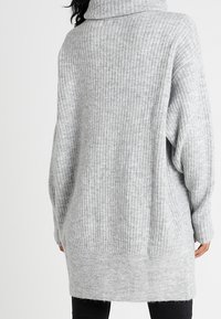 Harmaa, uritettu neulepaita, jossa on korkea kaulus, pitkät hihat ja rento istuvuus, joka ulottuu puoliväliin reisiä. Pehmeä tekstuuri, minimaalinen yksityiskohtaisuus.