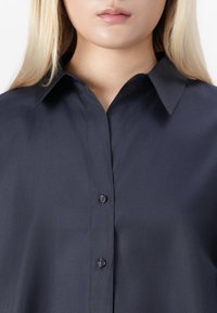 Woman with long blonde hair wearing a dark navy button-up shirt with a classic collar, visible from neck to mid-torso.