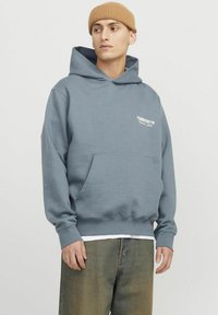 Sudadera gris con bolsillo frontal, puños acanalados y un logo en blanco. Combinada con unos vaqueros de cintura alta y un gorro beige.